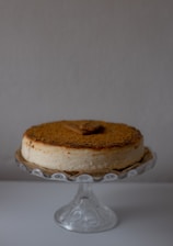 A creamy cheesecake with a crumbly brown topping is placed on a decorative glass stand. There is a rectangular cookie on top of the cheesecake, enhancing its presentation. The background is a plain, light color, which accentuates the cake as the focal point.