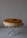 A creamy cheesecake with a crumbly brown topping is placed on a decorative glass stand. There is a rectangular cookie on top of the cheesecake, enhancing its presentation. The background is a plain, light color, which accentuates the cake as the focal point.