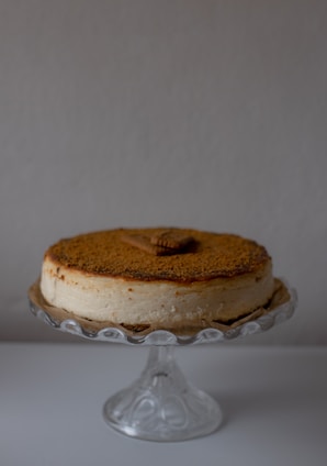 A creamy cheesecake with a crumbly brown topping is placed on a decorative glass stand. There is a rectangular cookie on top of the cheesecake, enhancing its presentation. The background is a plain, light color, which accentuates the cake as the focal point.