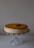 A creamy cheesecake with a crumbly brown topping is placed on a decorative glass stand. There is a rectangular cookie on top of the cheesecake, enhancing its presentation. The background is a plain, light color, which accentuates the cake as the focal point.