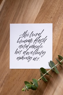 A piece of paper with elegant black calligraphy on a wooden surface, accompanied by a sprig of green eucalyptus.