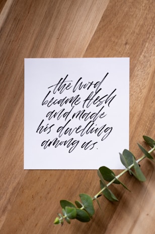 A piece of paper with elegant black calligraphy on a wooden surface, accompanied by a sprig of green eucalyptus.