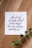 A piece of paper with elegant black calligraphy on a wooden surface, accompanied by a sprig of green eucalyptus.