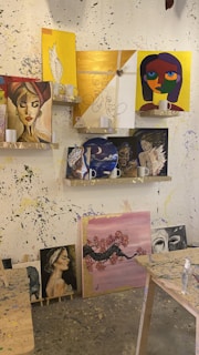 Various paintings are displayed on shelves and against a splattered wall. The paintings include portraits of women, abstract faces, and a cherry blossom tree. Some of the artworks are vivid and colorful, while others are more subdued. The shelves also hold white mugs. The room is an art studio with paint splatters on the wall and tables.