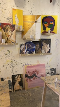 Various paintings are displayed on shelves and against a splattered wall. The paintings include portraits of women, abstract faces, and a cherry blossom tree. Some of the artworks are vivid and colorful, while others are more subdued. The shelves also hold white mugs. The room is an art studio with paint splatters on the wall and tables.