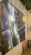 A painting inspired by Van Gogh's Starry Night with swirling blue, yellow, and white strokes and a bright crescent moon. A neon light outline of an abstract shape is superimposed on the painting. Additional artworks, including a painting of a woman's face with a green background, are leaning against the wall.