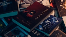A collection of books scattered on a surface, with one prominently featuring a dark cover with the title 'B&iacute; mật của Naoko' by Keigo Higashino. The books have a variety of covers with different colors and themes, including blue, black, and brown tones.