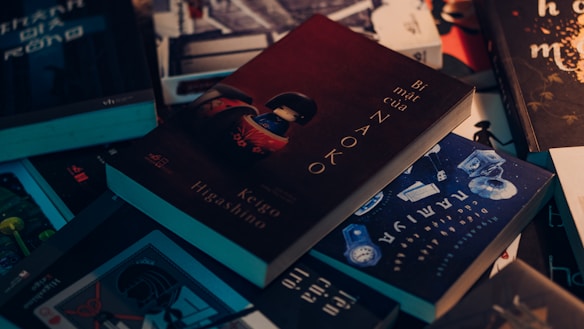 A collection of books scattered on a surface, with one prominently featuring a dark cover with the title 'B&iacute; mật của Naoko' by Keigo Higashino. The books have a variety of covers with different colors and themes, including blue, black, and brown tones.
