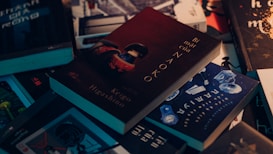 A collection of books scattered on a surface, with one prominently featuring a dark cover with the title 'Bí mật của Naoko' by Keigo Higashino. The books have a variety of covers with different colors and themes, including blue, black, and brown tones.