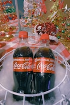 Two bottles of Coca-Cola with red caps are placed inside a white basket filled with white cotton-like material. Surrounding the basket are festive decorations, including gold and red ornaments and miniature Christmas trees, suggesting a holiday theme.