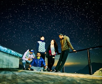 A group of five people are gathered on a rooftop or terrace with a starry night sky in the background. One person is seated on the ground with legs crossed, appearing to meditate. The others are standing or crouching nearby, engaged in conversation or enjoying the night view. The setting includes a metal railing and some mountainous terrain visible in the distance.