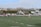 A group of people are playing rugby on a grass field. Several players are clustered around the ball, while others watch or prepare to engage. The players are wearing uniforms in black and white, and the referee is wearing a bright pink shirt. The field is surrounded by a hedge and a few benches, with a sparse audience watching from the sidelines.