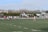 A group of people are playing rugby on a grass field. Several players are clustered around the ball, while others watch or prepare to engage. The players are wearing uniforms in black and white, and the referee is wearing a bright pink shirt. The field is surrounded by a hedge and a few benches, with a sparse audience watching from the sidelines.