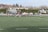 A group of athletes are playing rugby on a green field. The teams are distinguishable by their uniforms; one team wears black and white, while the other wears blue and black stripes. In the background, there are spectators sitting on steps and standing, watching the game. The setting is outdoors, possibly in a park or sports complex, with bare trees and bushes beyond the field.