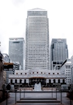 A cityscape featuring modern skyscrapers, with prominent buildings showcasing logos of major financial institutions. A fountain is placed in the foreground, surrounded by pedestrian walkways and railings.