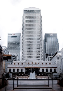 A cityscape featuring modern skyscrapers, with prominent buildings showcasing logos of major financial institutions. A fountain is placed in the foreground, surrounded by pedestrian walkways and railings.