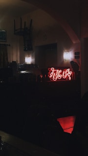 A dimly lit interior scene featuring a neon sign with Arabic script glowing red against the wall. Two wall lamps emit a soft light, adding to the atmospheric setting. There is an overturned chair hanging from the ceiling or wall, and a counter area with visible objects near the neon sign.