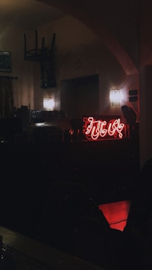 A dimly lit interior scene featuring a neon sign with Arabic script glowing red against the wall. Two wall lamps emit a soft light, adding to the atmospheric setting. There is an overturned chair hanging from the ceiling or wall, and a counter area with visible objects near the neon sign.
