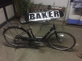 A black bicycle with a skateboard labeled 'BAKER' placed on its seat. The setting appears to be indoors or in a garage, with potted plants and storage containers in the background.