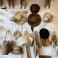A collection of hats and woven bags hangs on a wall. Various styles and colors, from straw hats to brown and black felt ones, showcase craftsmanship. The woven bags display textures and materials, including straw and canvas. Small hanging baskets and decorative elements add to the rustic aesthetic.