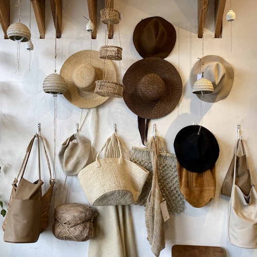 A collection of hats and woven bags hangs on a wall. Various styles and colors, from straw hats to brown and black felt ones, showcase craftsmanship. The woven bags display textures and materials, including straw and canvas. Small hanging baskets and decorative elements add to the rustic aesthetic.