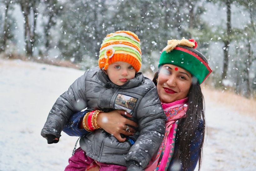 A woman and a child are outdoors in a snowy environment. The woman is wearing colorful traditional clothing and a hat, while the child is bundled up in a gray jacket and a vibrant orange and yellow striped beanie. They appear to be enjoying the snowfall, and the background is filled with trees covered in snow.
