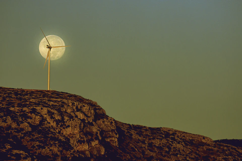 The image depicts a wind turbine standing tall on a rocky cliff, with its blades facing towards the right side of the frame and set against a backdrop of a full moon rising over the horizon. The sky above is painted in shades of greenish-blue, providing a serene contrast to the industrial nature of the wind turbine.