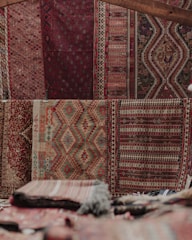An array of intricately patterned and colorful textile rugs displayed vertically on a wall, featuring geometric and symmetrical designs. The texture suggests traditional weaving techniques, with a mix of earthy and red tones. Stacks of folded rugs are visible in the foreground.
