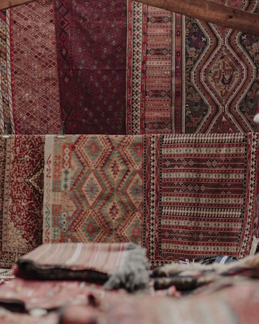 An array of intricately patterned and colorful textile rugs displayed vertically on a wall, featuring geometric and symmetrical designs. The texture suggests traditional weaving techniques, with a mix of earthy and red tones. Stacks of folded rugs are visible in the foreground.