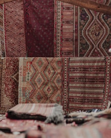 An array of intricately patterned and colorful textile rugs displayed vertically on a wall, featuring geometric and symmetrical designs. The texture suggests traditional weaving techniques, with a mix of earthy and red tones. Stacks of folded rugs are visible in the foreground.