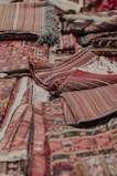 An assortment of intricately woven rugs with various patterns and textures is layered on top of each other. The rugs have a mix of geometric and linear designs, featuring rich colors such as reds, browns, and blacks. Fringes are visible on some of the edges, showcasing a traditional and handcrafted look.