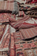 An assortment of intricately woven rugs with various patterns and textures is layered on top of each other. The rugs have a mix of geometric and linear designs, featuring rich colors such as reds, browns, and blacks. Fringes are visible on some of the edges, showcasing a traditional and handcrafted look.