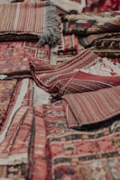 An assortment of intricately woven rugs with various patterns and textures is layered on top of each other. The rugs have a mix of geometric and linear designs, featuring rich colors such as reds, browns, and blacks. Fringes are visible on some of the edges, showcasing a traditional and handcrafted look.