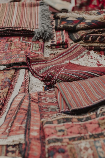 An assortment of intricately woven rugs with various patterns and textures is layered on top of each other. The rugs have a mix of geometric and linear designs, featuring rich colors such as reds, browns, and blacks. Fringes are visible on some of the edges, showcasing a traditional and handcrafted look.