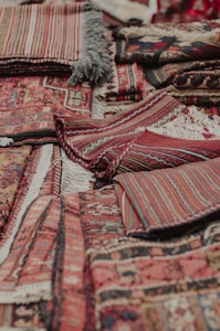 An assortment of intricately woven rugs with various patterns and textures is layered on top of each other. The rugs have a mix of geometric and linear designs, featuring rich colors such as reds, browns, and blacks. Fringes are visible on some of the edges, showcasing a traditional and handcrafted look.
