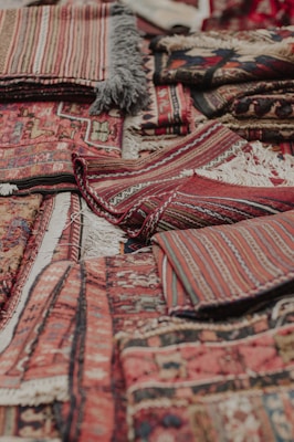 An assortment of intricately woven rugs with various patterns and textures is layered on top of each other. The rugs have a mix of geometric and linear designs, featuring rich colors such as reds, browns, and blacks. Fringes are visible on some of the edges, showcasing a traditional and handcrafted look.