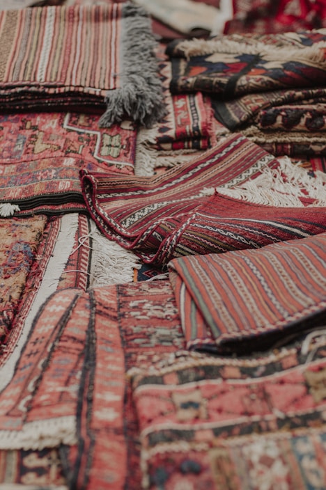 An assortment of intricately woven rugs with various patterns and textures is layered on top of each other. The rugs have a mix of geometric and linear designs, featuring rich colors such as reds, browns, and blacks. Fringes are visible on some of the edges, showcasing a traditional and handcrafted look.