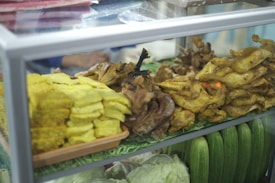 A display case contains various food items including yellow tofu and several portions of cooked poultry. Arranged alongside them are fresh green cucumbers and sliced cabbage. The setting appears to be a market or food stall.