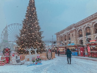 A festive outdoor scene in the wintertime, featuring a large Christmas tree adorned with lights and ornaments. There is a snowy setting with decorated stalls along the side, indicating a holiday market. A person dressed warmly is walking through the snow. In the background, a Ferris wheel and an ornate building with columns can be seen.