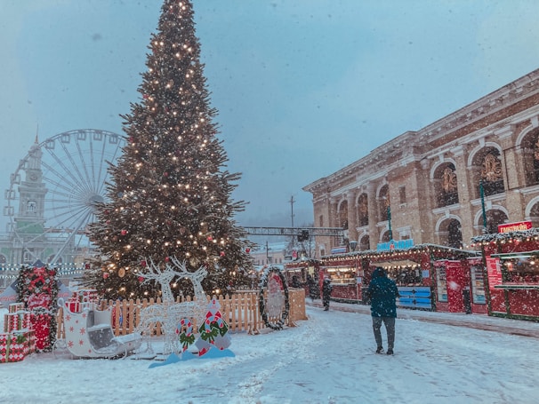A festive outdoor scene in the wintertime, featuring a large Christmas tree adorned with lights and ornaments. There is a snowy setting with decorated stalls along the side, indicating a holiday market. A person dressed warmly is walking through the snow. In the background, a Ferris wheel and an ornate building with columns can be seen.