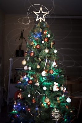 A beautifully decorated Christmas tree adorned with colorful lights and ornaments, featuring a prominent star at the top. Surrounding the tree are various round, sparkling ornaments in gold, red, and white colors. The tree is lush and dark green, with soft, swirling, blurred lights that create a magical, festive atmosphere.