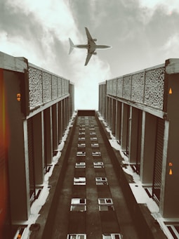 A tall, symmetrical building with intricate patterned fa&ccedil;ades on either side stretches upwards, leading the viewer's eye toward the sky. A commercial airplane is captured in mid-flight, centered against a backdrop of cloud-filled sky, creating a dynamic contrast between architecture and aviation.