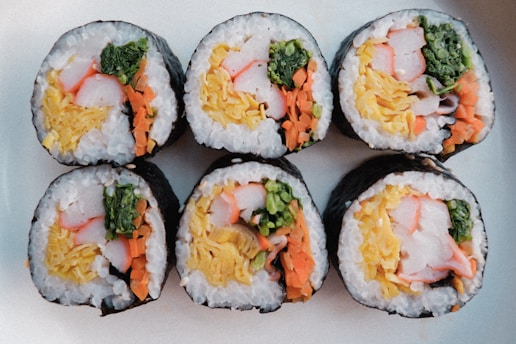 Six pieces of sushi rolls are neatly arranged on a light-colored surface. Each roll is filled with ingredients like crab stick, leafy greens, shredded carrots, and rice, all wrapped in a seaweed sheet.
