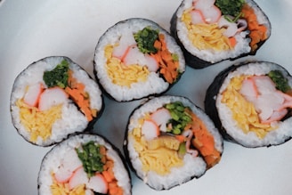 Several pieces of sushi rolls arranged on a white plate. The rolls contain ingredients like crab sticks, shredded egg omelet, carrot, and leafy greens wrapped in rice and seaweed.