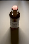 A glass bottle with a labeled exterior and a red top, casting a shadow on a light-colored surface. The label includes the brand name and product details, and the lighting gives the bottle a reflective sheen.