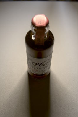 A glass bottle with a labeled exterior and a red top, casting a shadow on a light-colored surface. The label includes the brand name and product details, and the lighting gives the bottle a reflective sheen.