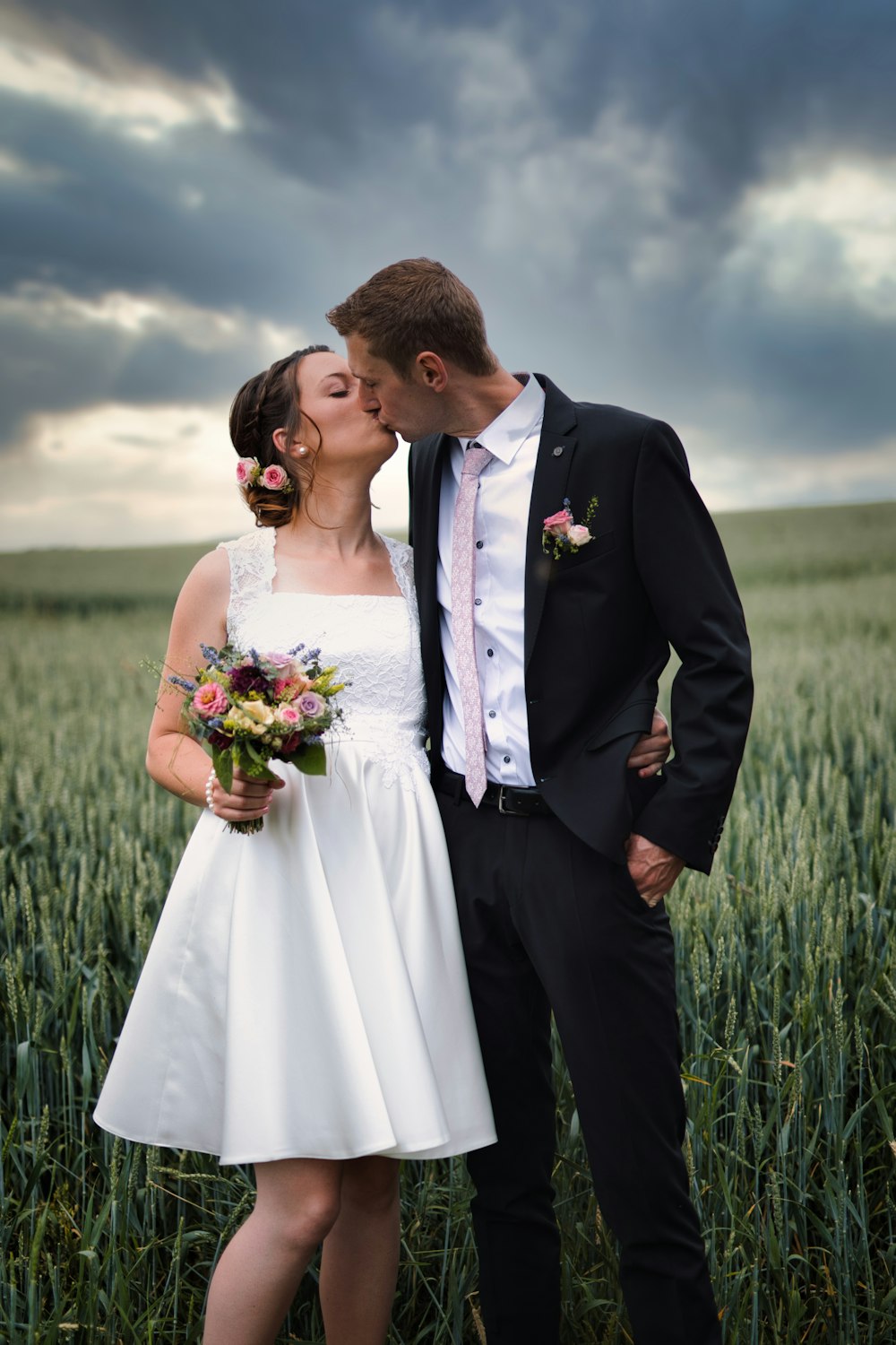 man in black suit kissing woman in white wedding dress on green grass field during daytime