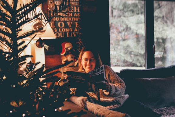 A cozy indoor scene featuring a woman wearing glasses and a knitted sweater, reading a book by a decorated Christmas tree. Warm lights illuminate the room, enhancing the festive and inviting atmosphere. Outside, snow-covered trees are visible through the window, adding to the wintry ambiance.