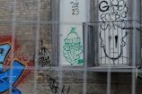 The image features a section of a brick wall covered with various graffiti designs. A vibrant piece of street art with blue and red hues is present on the left side. In the center, there's a green graffiti illustration of a character wearing glasses and a winter hat holding a pipe. On the right, a black outline of a stylized face with bold writing is visible through some metal bars.
