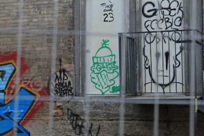 The image features a section of a brick wall covered with various graffiti designs. A vibrant piece of street art with blue and red hues is present on the left side. In the center, there's a green graffiti illustration of a character wearing glasses and a winter hat holding a pipe. On the right, a black outline of a stylized face with bold writing is visible through some metal bars.